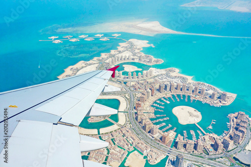 Aerial view from airplane window over The Pearl-Qatar artificial island in Doha, Qatar, with aircraft wing visible above turquoise waters, luxury marinas and modern waterfront architecture during flig