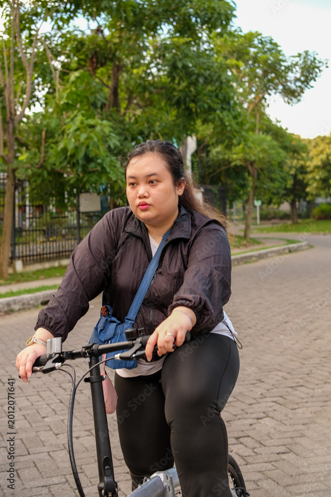 Obraz premium Woman riding a bicycle outdoors, looking thoughtful, with trees and a paved path in the background.
