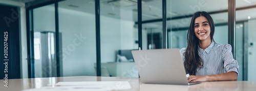 Smiling woman working on a laptop at an office desk with glass walls