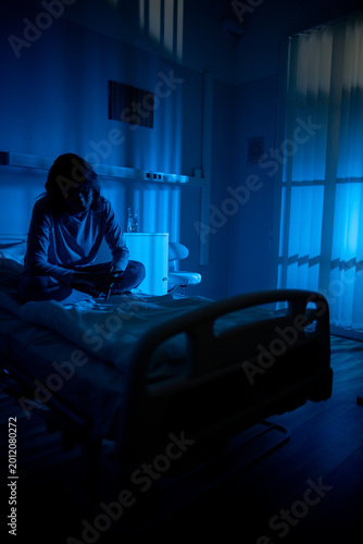 Patient sitting in dim hospital room, silhouette facing empty bed and window light while waiting for next steps mood balances hope and uncertainty in latenight clinical setting