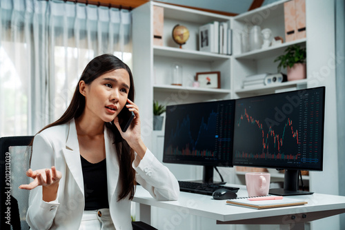 Tableau sur toile Stressful young beautiful Asian businesswoman calling to broker on smartphone, analyze possible risk to profit or loss in dynamic exchange stock market graph on pc screen at modern office