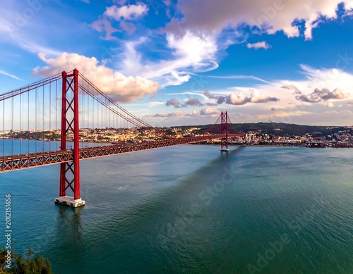A scenic view of a red suspension bridge over tranquil blue waters. City backdrop, sunny sky