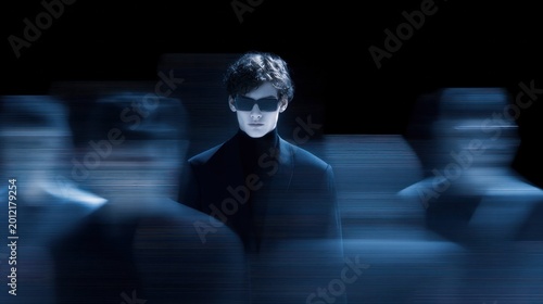 Man wearing sunglasses and black turtleneck stands still with blurred figures moving around him, concept of isolation, focus, and contrast in dark environment with motion effect.