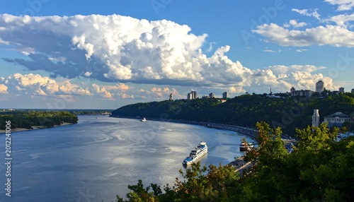 A scenic vista of a broad river flowing past a green hillside, city skyline on the right. Bright blue skies, puffy clouds