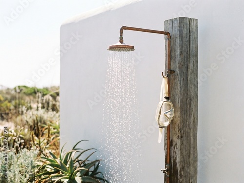Rustic outdoor shower with a copper-toned head and wooden post spraying water on a sunny day, set against a white wall and natural landscape.