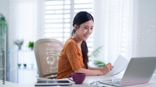 Asian woman working from home office writing document reading paperwork using laptop computer sitting desk bright modern workspace remote hybrid work freelance