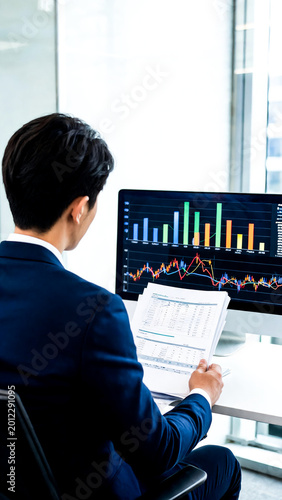 Asian Businessman Sitting at Office Desk Reviewing Financial Report Documents While Analyzing Multiple Colorful Charts and Graphs on Computer Monitor for Investment Strategy Decision