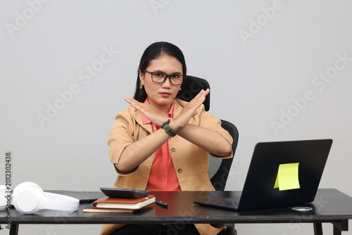 A young woman with a serious expression, working sitting at an office desk while making a gesture of crossing her arms as a sign of stopping or prohibition.
