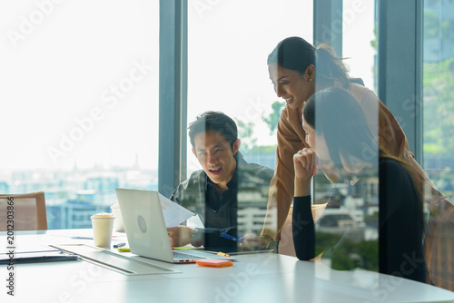 Tableau sur toile A group of multi-ethnic young professionals, aged 20-30, working together around a laptop in a bright office with large windows
