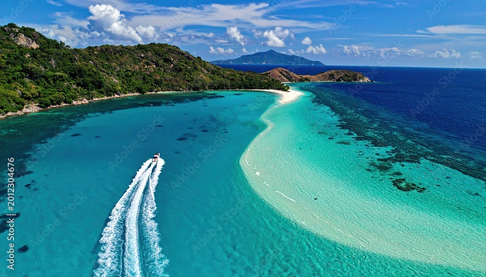 Obraz premium Aerial view of a speedboat cruising through turquoise water near a tropical island sandbar.