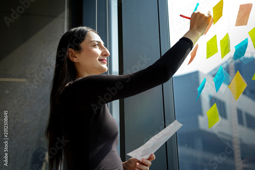 A focused young professional woman planning and organizing ideas using vibrant sticky notes on a glass window. The images capture a creative workflow, project management, and strategy development in a