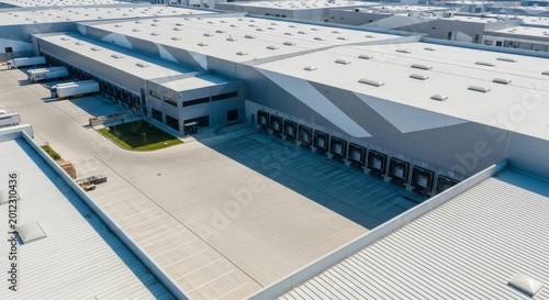 A high-angle drone perspective of a modern industrial warehouse complex featuring loading docks, vast rooftops, and a gray asphalt yard, architecture, logistics center, roof