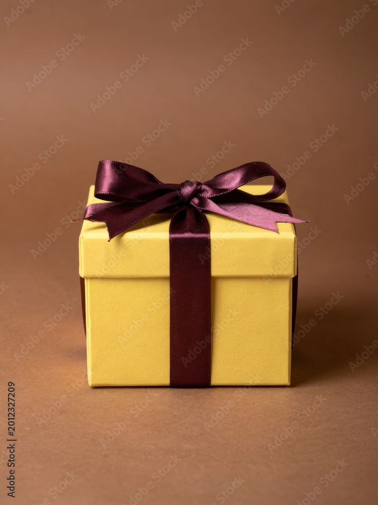 Fototapeta premium Yellow square gift box with a purple satin ribbon bow tied on top sits on a brown surface for holiday gifting, seasonal retail campaigns and festive celebrations