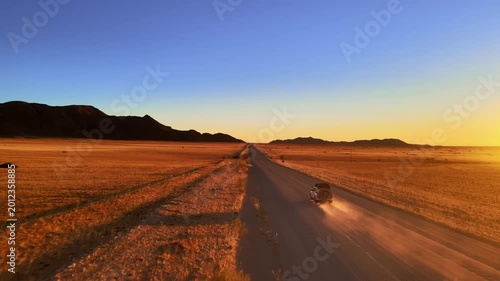 A jeep drives along a dusty road through the Namib Desert at sunset, filmed by drone, surrounded by glowing red dunes and golden light, capturing the wild beauty and freedom of desert adventure.
