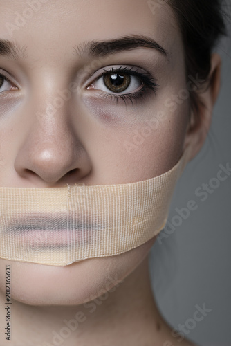 Close-up portrait of a young woman with her mouth covered by medical tape, symbolizing censorship, domestic violence, being silenced, and the struggle for freedom of speech in a modern world