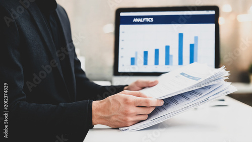 Businessman Holding Stack of Financial Report Documents While Reviewing Analytics Bar Chart Data on Computer Screen at Modern Office for Business Decision Making