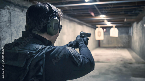 A man wearing protective headphones and tactical gear is practicing pistol shooting in a closed range. A back view of the shooter aiming at paper targets.