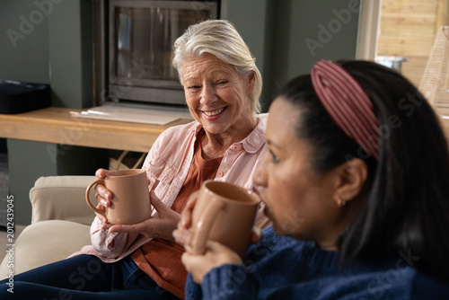 Senior and mature female friends smiling and sipping tan mugs on beige sofa in living room