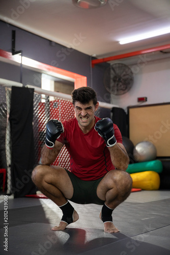 Mid-adult man squatting on padded mat at martial arts gym, wearing red shirt, gloves, ankle wraps