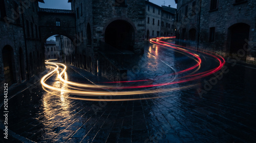 Dynamic light trails streak through a historic European cobblestone street at dusk, reflecting on wet surfaces