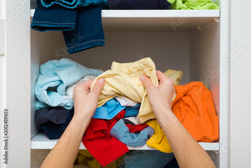Mother hands sorting cluttered different colorful clothes on shelves in white opened wooden wardrobe at home room. Messy casual kid apparel. Making neatness. Closeup. Point of view shot. Front view.