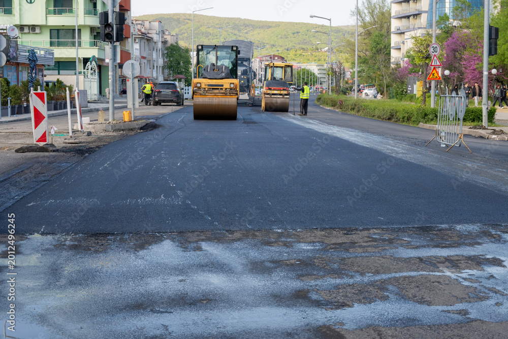 Fototapeta premium Two road rollers work in tandem to flatten a wide stretch of fresh, dark asphalt on a city boulevard during an extensive road renovation project.