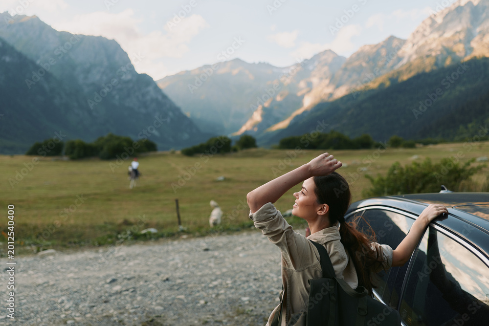Naklejka premium Woman beside a car in a mountain meadow, enjoying fresh air and wide landscape, conveys travel freedom and outdoor lifestyle in a sunny rural scene