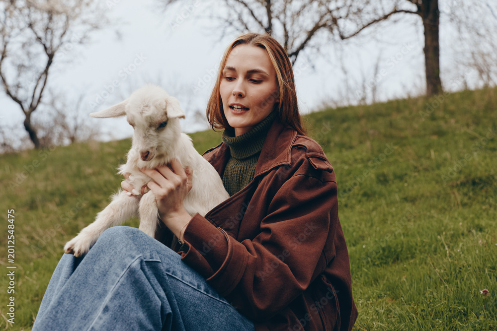 Obraz premium Cheerful woman outdoors, seated on grass, holding a young goat kid, warm jacket, gentle breeze, sunlit scene, natural colors, field background, candid lifestyle moment, calm connection with animals
