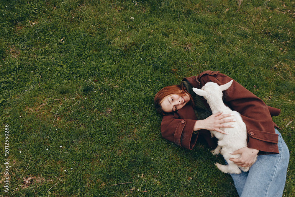 Obraz premium Young woman with red hair lies on green grass holding a white baby goat and smiling happily in a relaxed outdoor lifestyle scene.