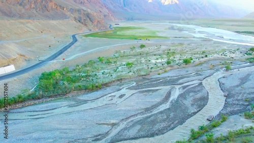 Wide panoramic landscape of a mountain river delta in a barren wilderness with green oasis patches and steep slopes.

