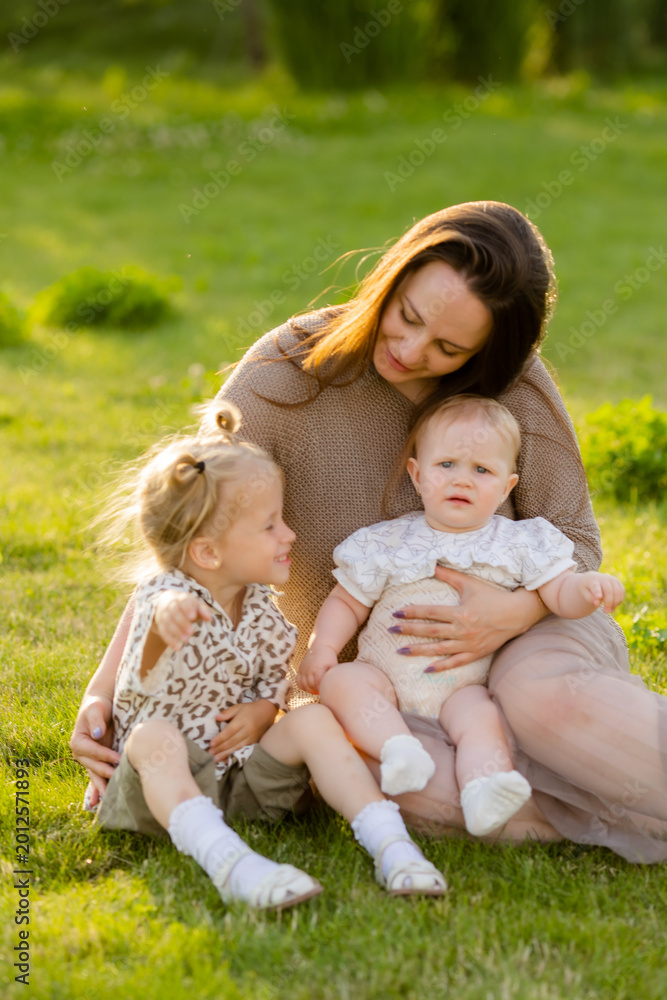 Fototapeta premium mother with two daughters sitting on a lawn at sunset in the summer, hugging