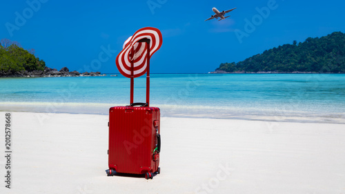 Summer vacation concept with red suitcase and sun hat on a tropical white sand beach with an airplane flying above and Travel tropical getaway background