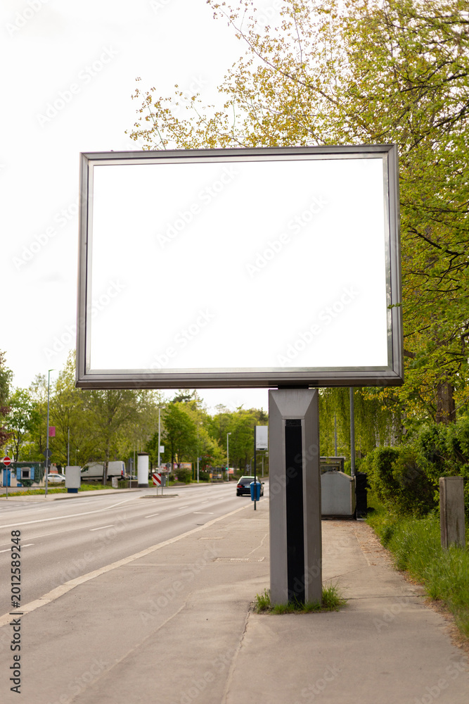 Fototapeta premium A large empty billboard is positioned next to a road. The surrounding area features green trees and minimal traffic. It is a clear day with sunlight