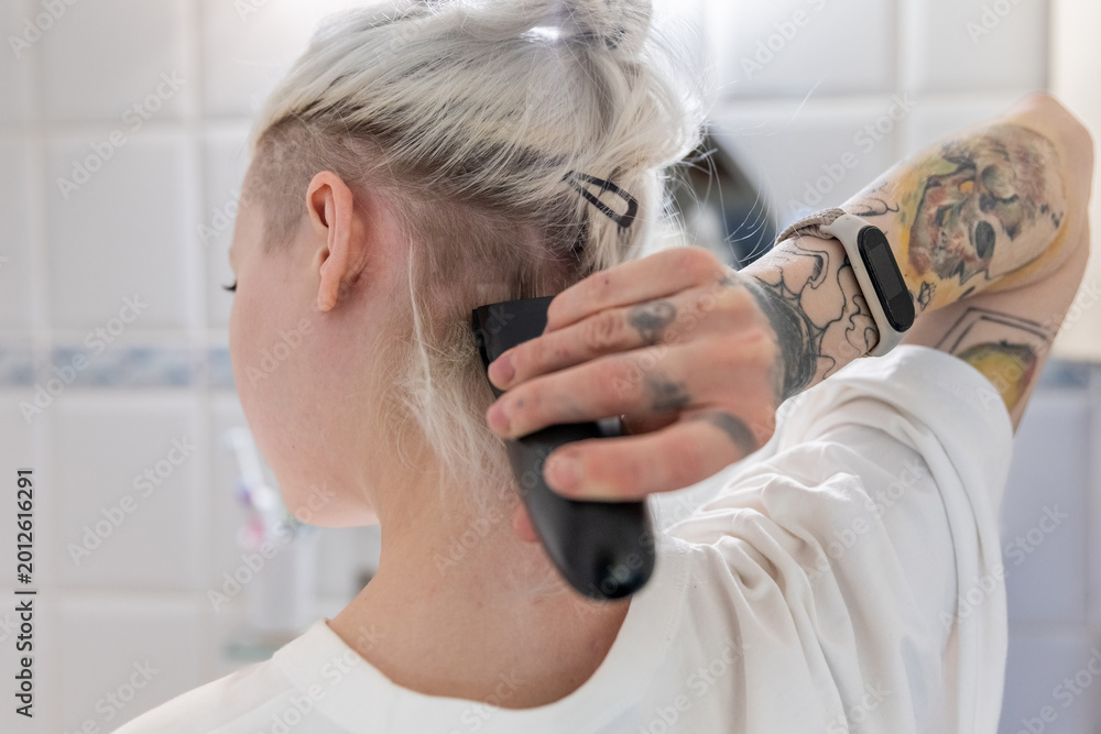 Fototapeta premium A young woman with tattoos and piercings shaving back of her head in the bathroom
