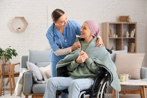 Mature woman after chemotherapy in wheelchair with warm plaid and nurse at ho...