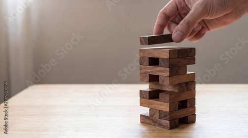 Photos Hand carefully balancing a wooden block atop a tall, precarious tower, representing risk management, precision