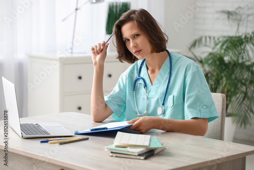 Beautiful female doctor with clipboard and modern laptop at table in clinic