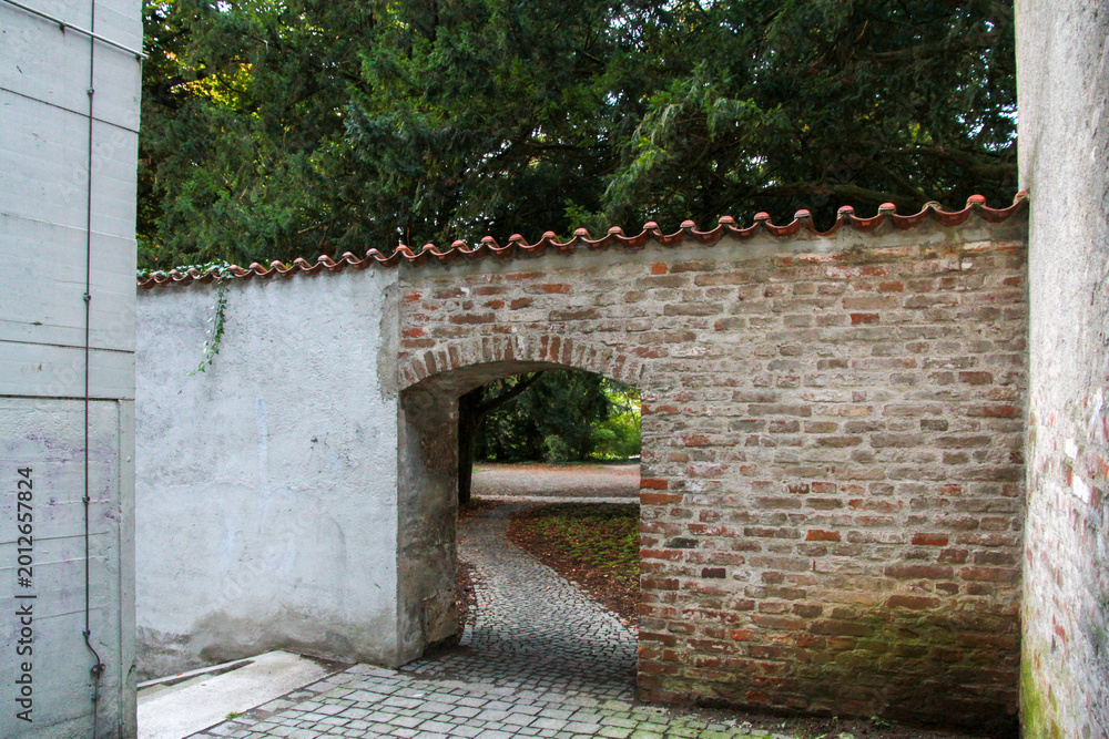 Obraz premium Ancient brick wall with an arched gateway leading to a lush green park. Traditional European street architecture with cobblestone path and historic masonry in Bavaria, Germany.