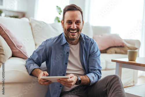 Bild auf Leinwand Smiling Man Using Tablet At Home In Bright Living Room, Casual Remote Work And R