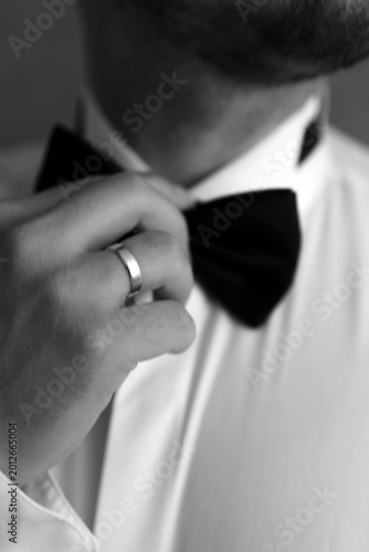 Black and white close-up of a man adjusting his bow tie, wearing a formal white dress shirt and a wedding ring, preparing for a gala, celebration, or official ceremony.