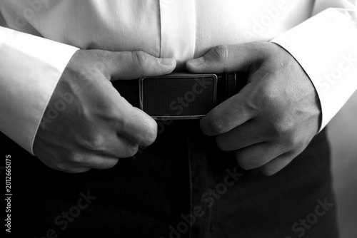 Photography Black and white close-up of a man's hands fastening a leather belt with a metal buckle on formal trousers