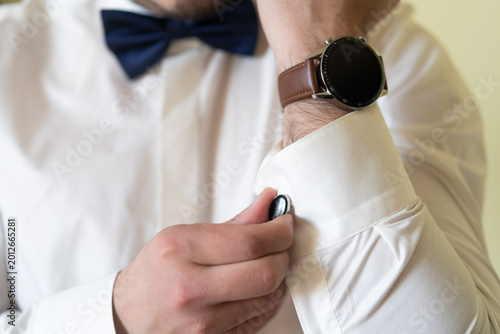 Detail shot of a man in a white dress shirt and navy blue bow tie fastening a black cufflink. Featuring a stylish wristwatch with a brown leather strap for a formal event look.
