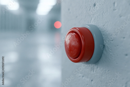 Red button glows softly on gray wall. Faint bokeh lights blur in background. Button seems ready to be pressed. Suggests control, action, or decision. Ideal for themes of choice or activation