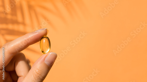Close-up of hand holding golden softgel capsule with tropical shadows, premium wellness and oral sunscreen concept on apricot orange background with copy space.