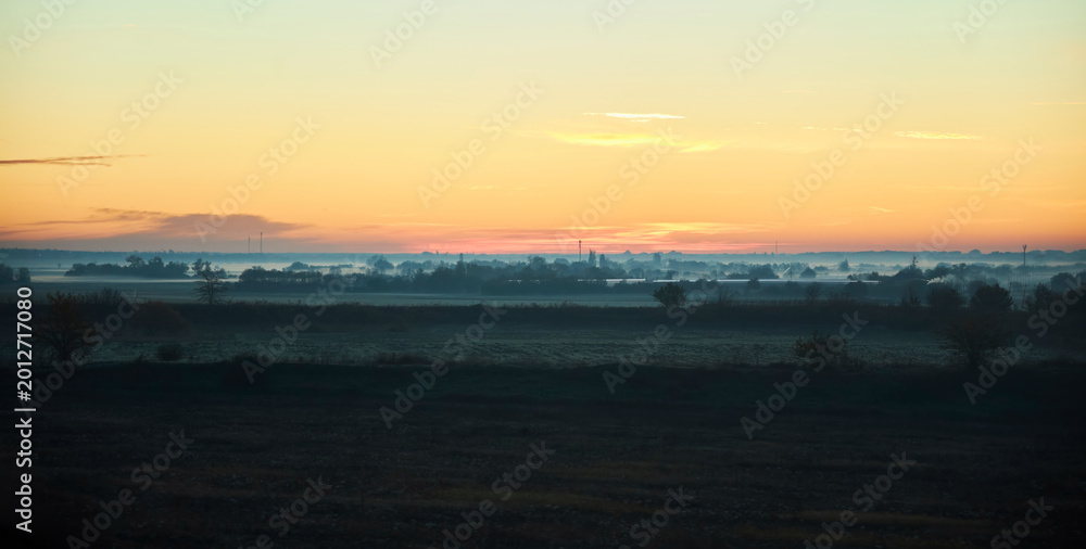 Obraz premium A misty dawn envelops the rolling fields and distant trees. The twilight sky is painted in soft pink and orange tones. Fog envelops the grass