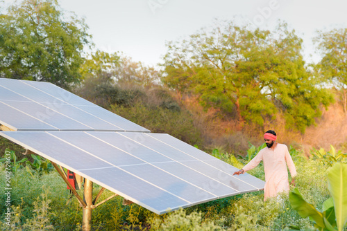A confident indian farmer or rural worker stands in front of solar panels at agriculture field