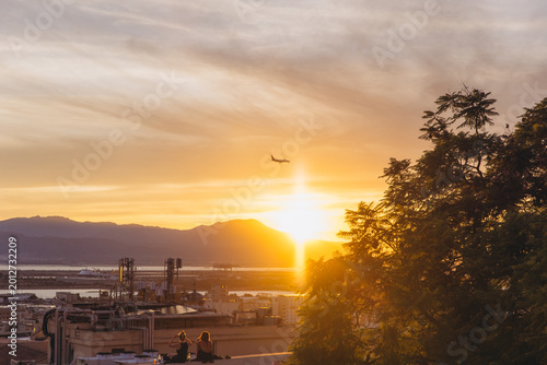 Tableau sur toile Women relaxing on rooftop watching airplane flying over Cagliari at sunset, cont
