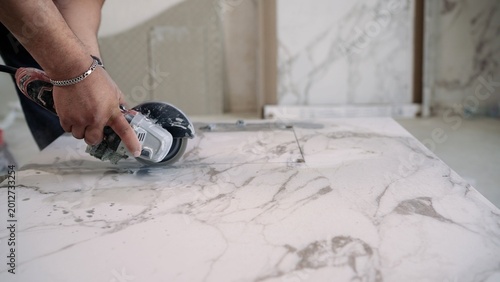 A construction worker uses an angle grinder to cut a large white marble or porcelain tile. Close up shot of hands guiding power tool along a straight line for a precise cut during renovation