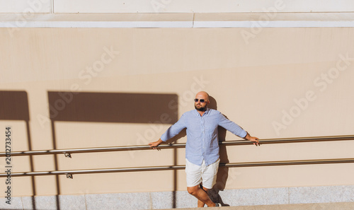 Fotografía Man leaning on a railing against a beige wall, enjoying the sunny urban travel e