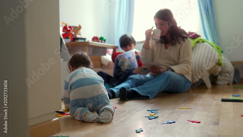 Foto Mother sitting on floor with children, holding up toy while baby crawls nearby a